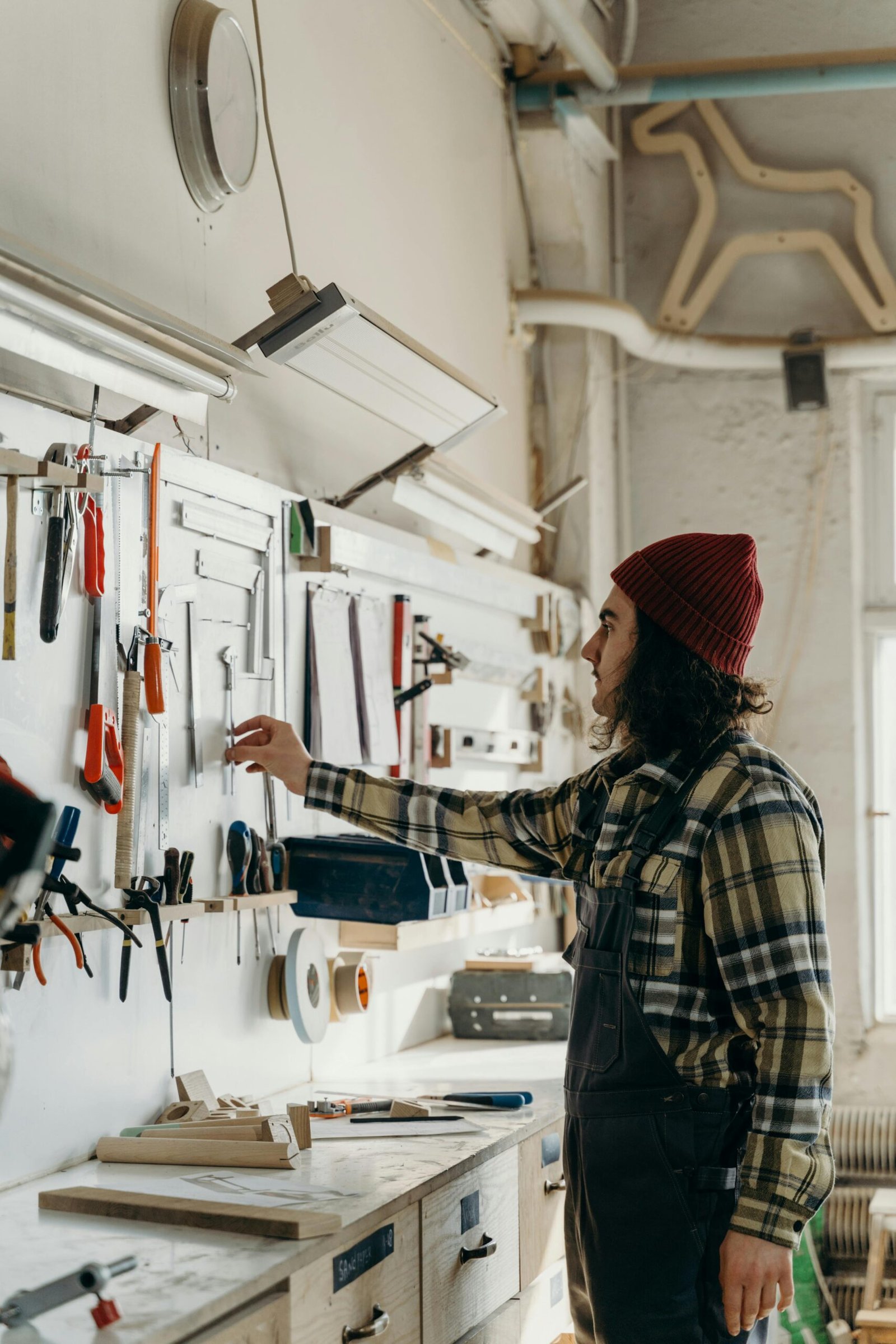 pexels-photo-7480722-7480722 A craftsman in a beanie selecting tools from a workshop wall with organized equipment.